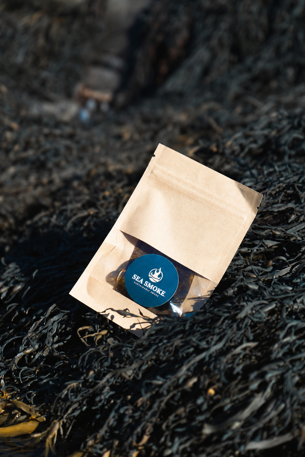 Sea Smoke kraft pouch with a blue circular label and clear window, resting among dark coastal seaweed.